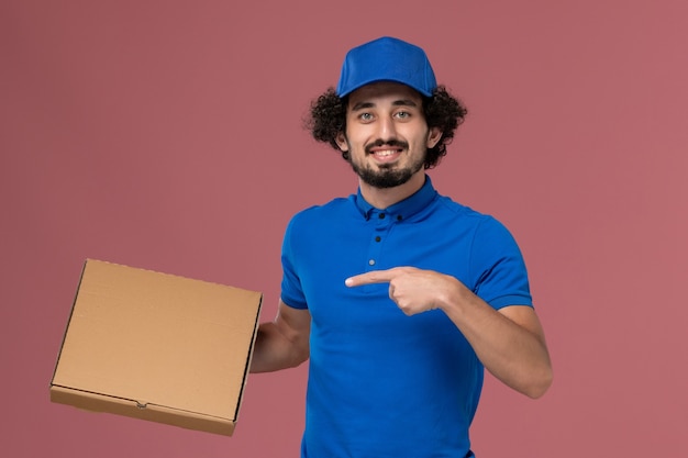 Front view of male courier in blue uniform cap with food box on his hands on light-pink wall