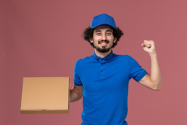 Front view of male courier in blue uniform cap with food box on his hands on light-pink wall