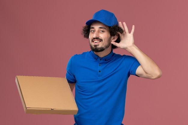 Front view of male courier in blue uniform cap with food box on his hands on light-pink wall