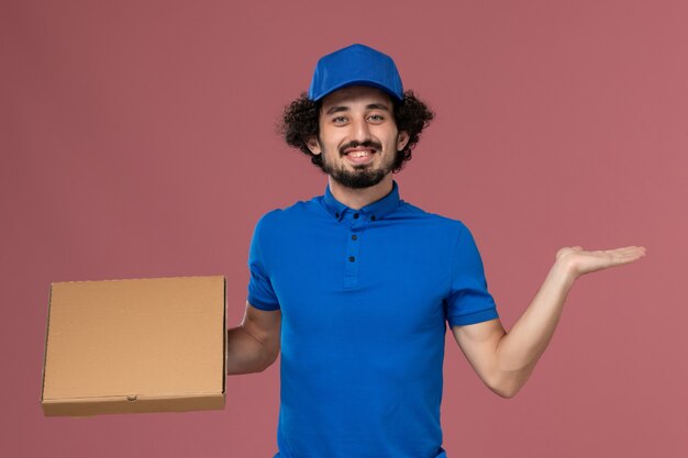 Вид спереди курьера-мужчины в синей форменной кепке с коробкой для еды на руках на светло-розовой стене