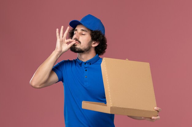 Front view of male courier in blue uniform cap with food box on his hands opening it on light-pink wall