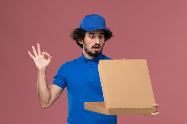 밝은 분홍색 벽에 그것을 여는 그의 손에 음식 상자와 파란색 유니폼 모자에 남성 택배의 전면보기