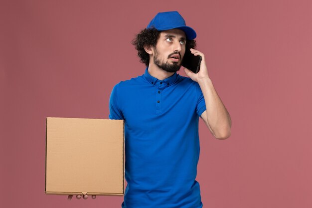 Front view of male courier in blue uniform cap with food box on his hands talking on the phone on the light-pink wall