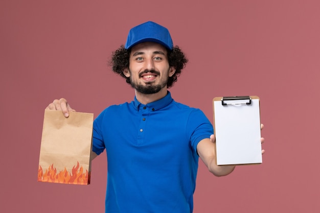 Vista frontale del corriere maschio in protezione uniforme blu con blocco note e pacchetto di cibo di consegna sulle sue mani sul muro rosa chiaro