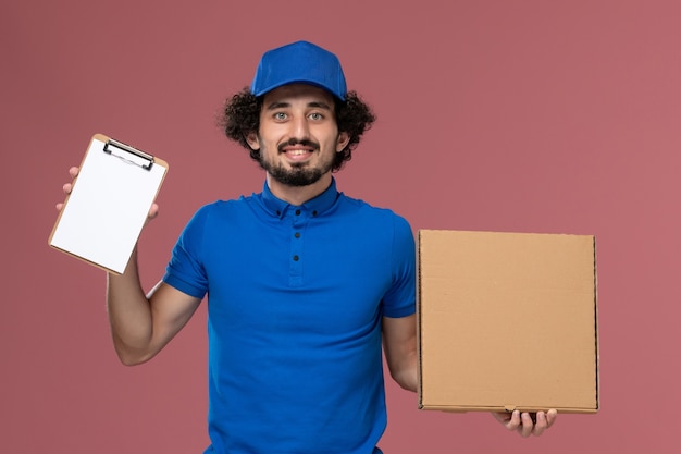 Free photo front view of male courier in blue uniform cap with notepad and food box on his hands on pink wall