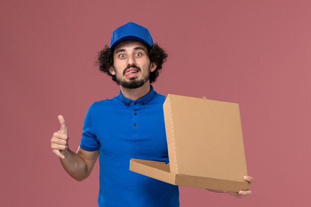 Front view of male courier in blue uniform cap with open food box on his hands on light-pink wall