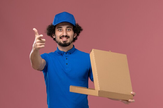 Front view of male courier in blue uniform cap with open food box on his hands on the light-pink wall