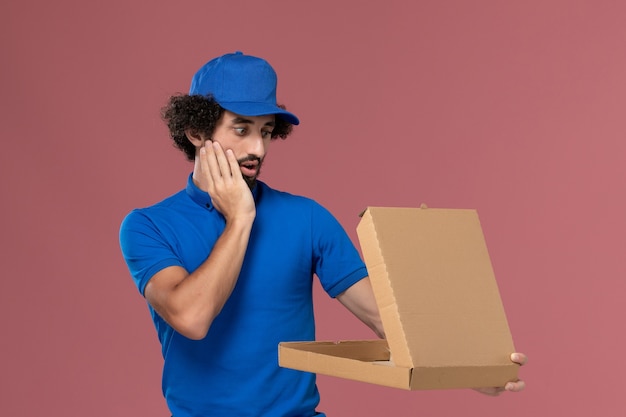 밝은 분홍색 벽에 그의 손에 열린 음식 상자가있는 파란색 유니폼 모자 남성 택배의 전면보기