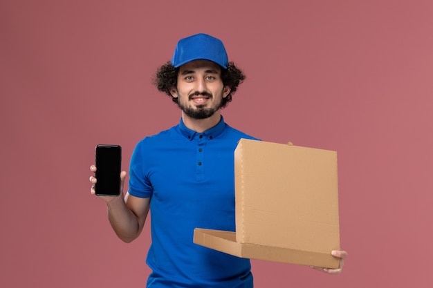 Front view of male courier in blue uniform cap with open food box and phone on his hands on the light-pink wall