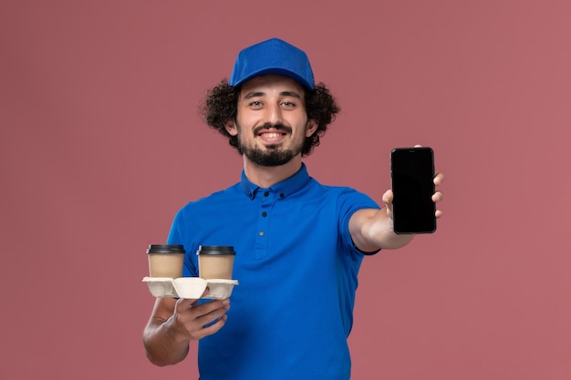 Front view of male courier in blue uniform and cap with phone and delivery coffee cups on his hands on pink wall