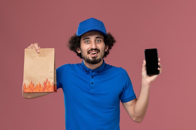 Front view of male courier in blue uniform cap with smartphone and delivery food package on his hands on the pink wall