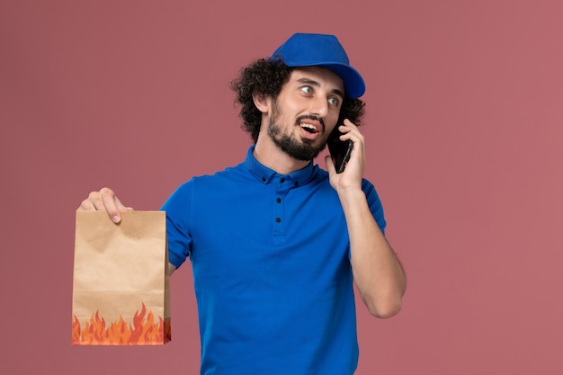 분홍색 벽에 그의 손에 스마트 폰 및 배달 음식 패키지와 함께 파란색 유니폼 모자에 남성 택배의 전면보기