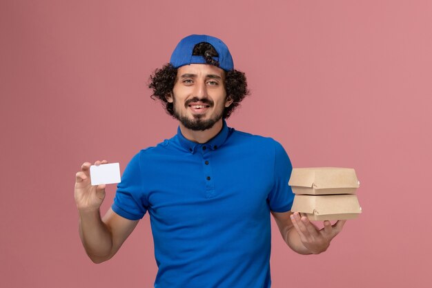 Front view male courier in blue uniform and cape holding delivery food packages with card on the pink wall uniform delivery male service