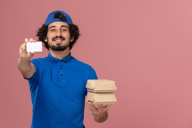 青いユニフォームとピンクの壁にカード付きの配達食品パッケージを保持しているケープの正面図男性宅配便制服配達サービス男性