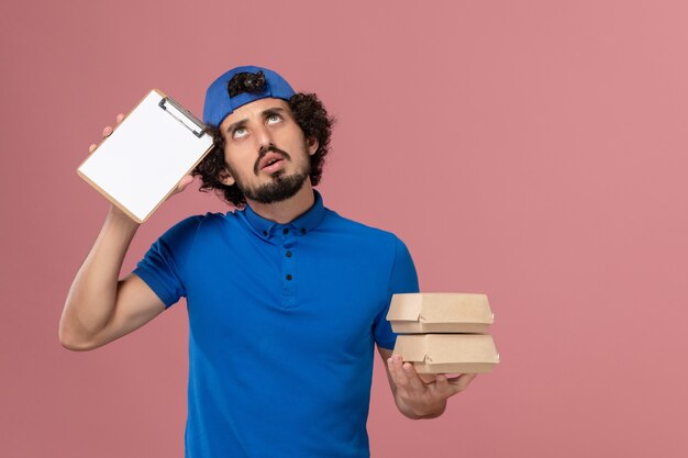 Front view male courier in blue uniform and cape holding delivery food packages with notepad on pink wall uniform delivery service job