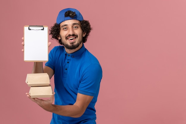 Foto gratuita corriere maschio di vista frontale in uniforme blu e mantello che tiene i pacchetti di cibo di consegna con il blocco note sul servizio di consegna uniforme del lavoro della parete rosa