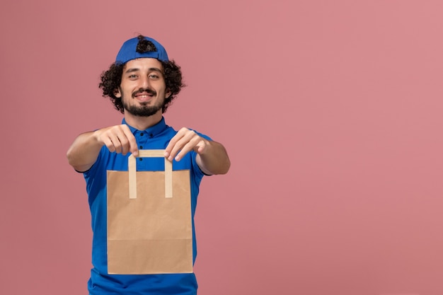 Corriere maschio di vista frontale in uniforme blu e mantello che tiene il pacchetto di cibo di carta di consegna sulla uniforme di lavoro di servizio di consegna della parete rosa