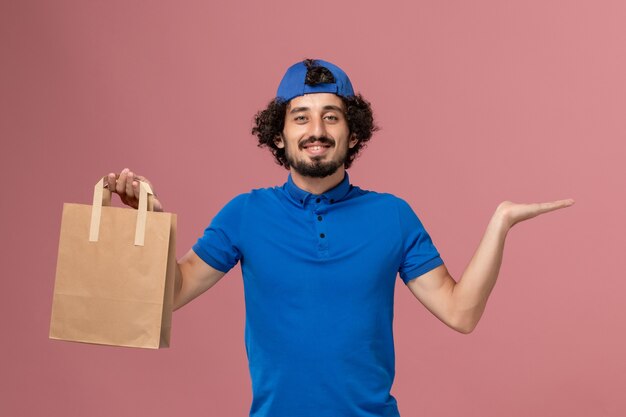 Front view male courier in blue uniform and cape holding delivery paper food package on pink wall delivery service uniform work