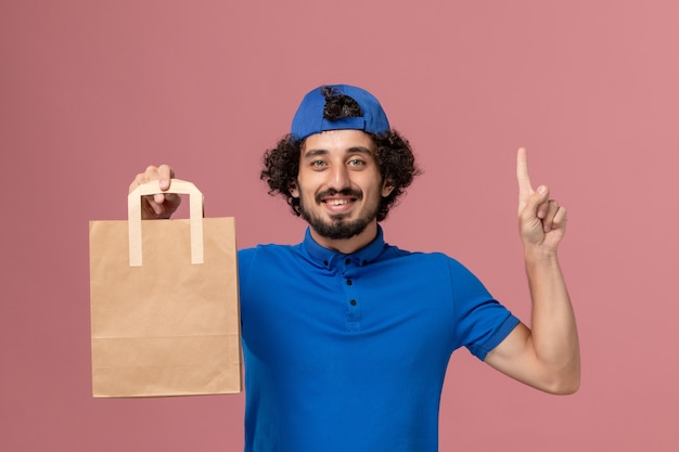 Corriere maschio di vista frontale in uniforme blu e mantello che tiene il pacchetto di cibo di carta di consegna sull'uniforme di servizio di consegna della parete rosa