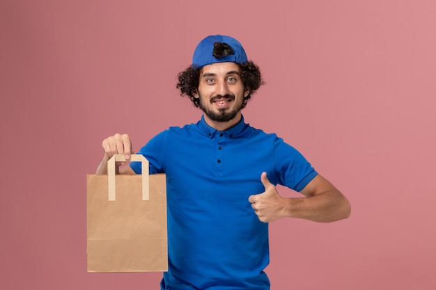 파란색 유니폼과 케이프 핑크 벽 작업 배달 서비스 유니폼에 배달 종이 음식 패키지를 들고 전면보기 남성 택배