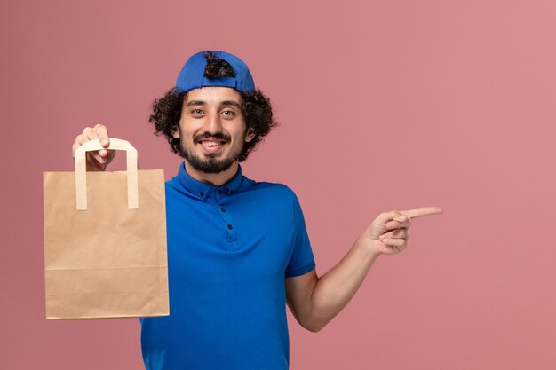 Front view male courier in blue uniform and cape holding delivery paper food package on pink wall worker male delivery service uniform