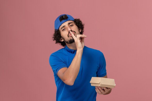 Front view male courier in blue uniform and cape holding little delivery food package on pink wall uniform delivery service company