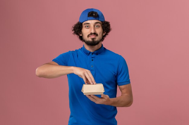 Front view male courier in blue uniform and cape holding little delivery food package on pink wall