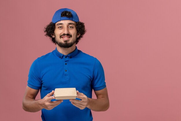 Front view male courier in blue uniform and cape holding little delivery food package on pink wall