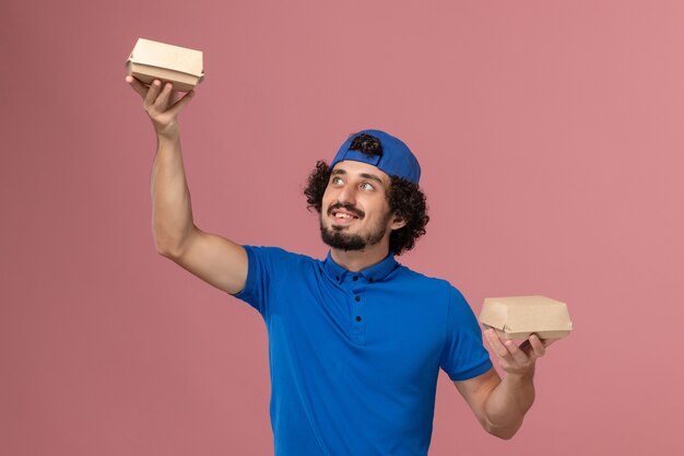 Front view male courier in blue uniform and cape holding little delivery food packages on the pink wall