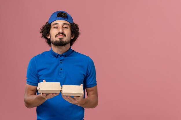 Front view male courier in blue uniform and cape holding little delivery food packages on the pink wall