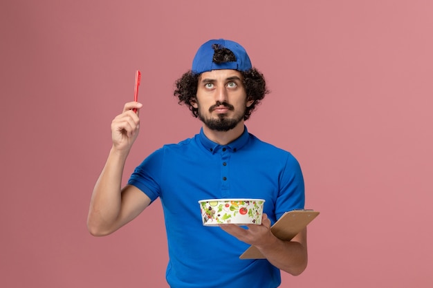 Курьер-мужчина, вид спереди в синей униформе и плаще, держит ручку для блокнота и круглую миску для доставки на розовой стене