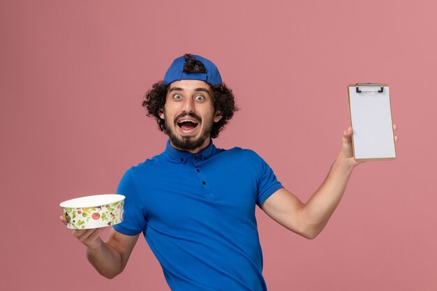 Front view male courier in blue uniform and cape holding notepad and round delivery bowl on pink wall