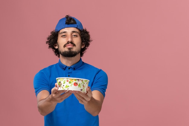 Front view male courier in blue uniform and cape holding round delivery bowl on pink wall