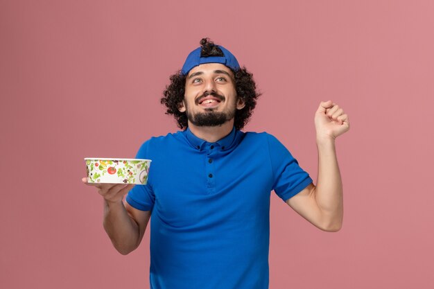 Front view male courier in blue uniform and cape holding round delivery bowl on pink wall