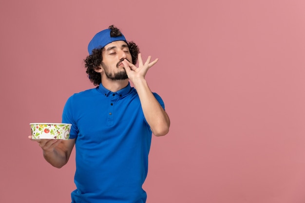 Front view male courier in blue uniform and cape holding round delivery bowl on pink wall