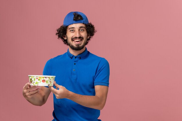 Front view male courier in blue uniform and cape holding round delivery bowl on the pink wall