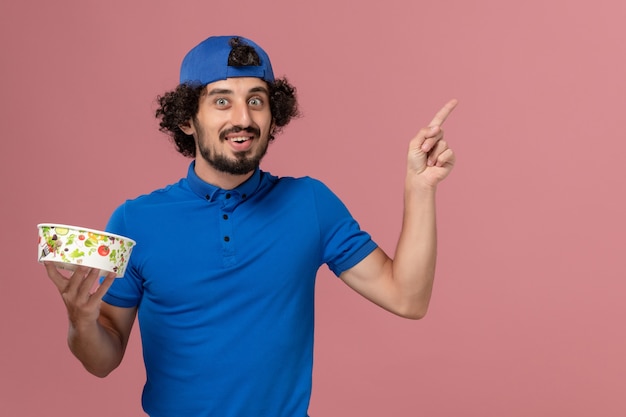 Front view male courier in blue uniform and cape holding round delivery bowl on the pink wall