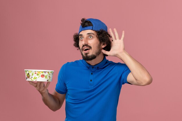 Front view male courier in blue uniform and cape holding round delivery bowl trying to hear on the pink wall