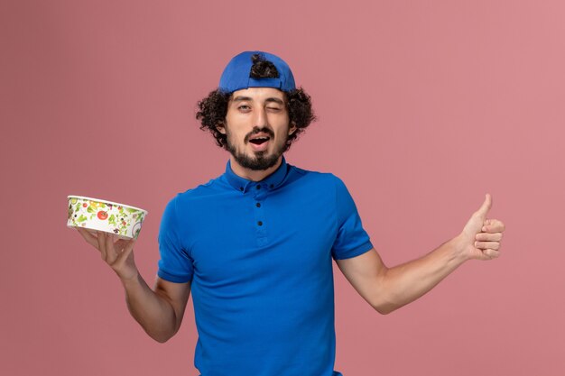 Front view male courier in blue uniform and cape holding round delivery bowl and winking on the pink wall