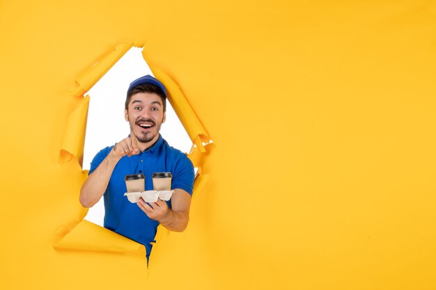 Front view male courier in blue uniform holding coffee cups on the yellow space