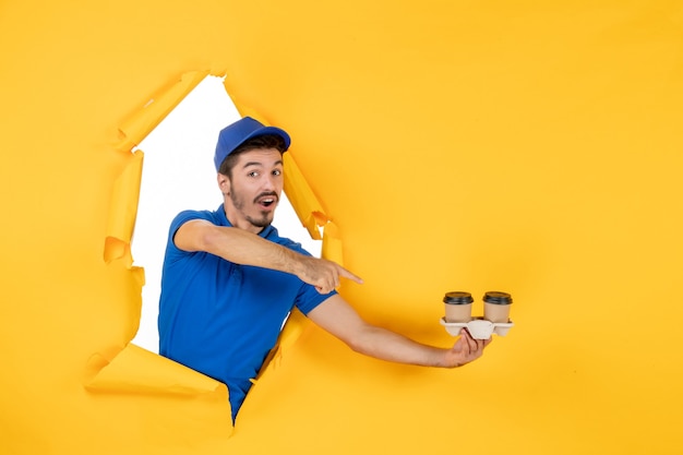 Front view male courier in blue uniform holding coffee cups on yellow space