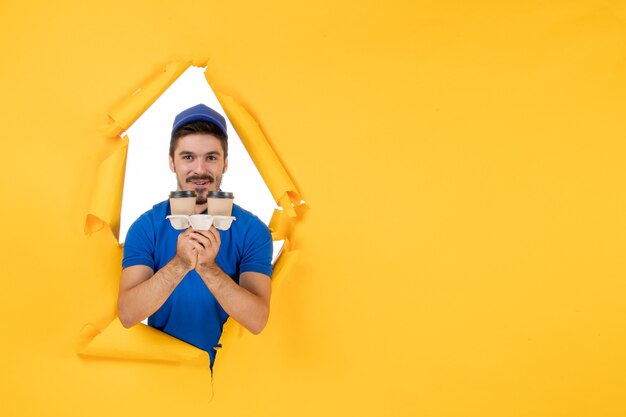 Front view male courier in blue uniform holding coffee cups on yellow space