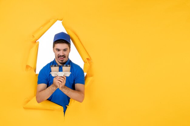 Front view male courier in blue uniform holding coffee cups on yellow space