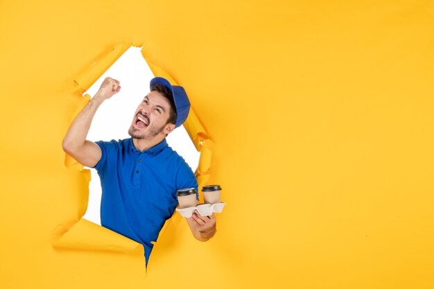 Front view male courier in blue uniform holding coffee cups on yellow space