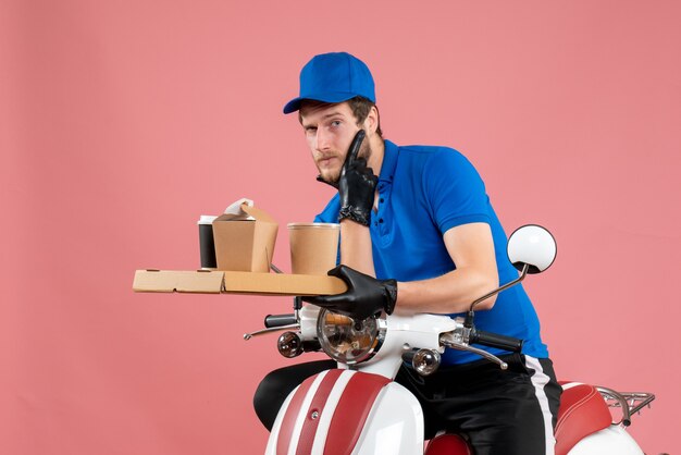 Front view male courier in blue uniform holding coffee and food box on the pink service fast-food work delivery job bike color