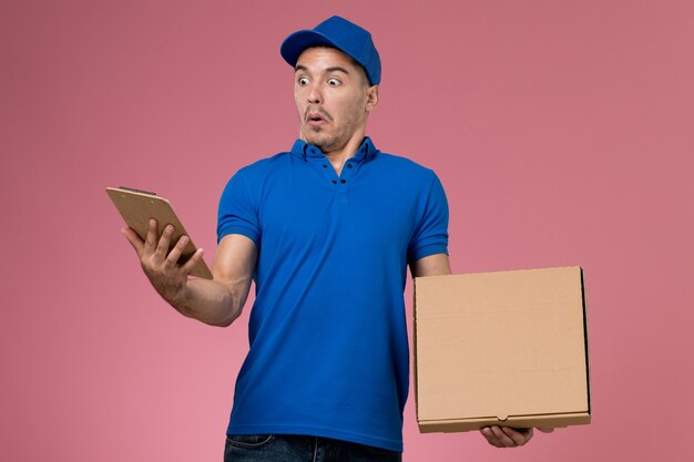 Курьер-мужчина, вид спереди в синей форме, держит коробку для еды с блокнотом на розовой стене, служба доставки униформы