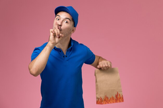 Front view male courier in blue uniform holding food package asking to be silent on pink wall, job worker uniform service delivery