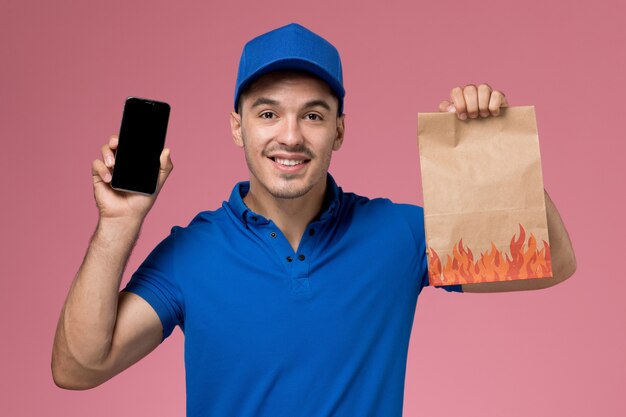 ピンクの壁に食品パッケージと電話を保持している青い制服の正面図男性宅配便、均一なサービス提供