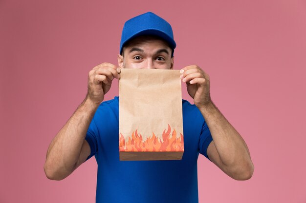 Front view male courier in blue uniform holding food package on pink wall, job worker uniform service delivery