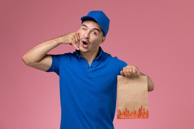 Front view male courier in blue uniform holding food package on the pink wall, job worker uniform service delivery